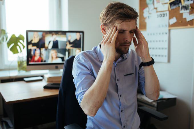 A man rubbing his head in stress with a Zoom meeting happening behind him