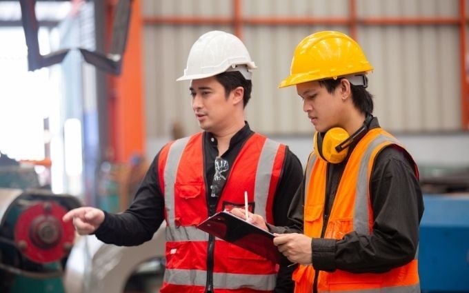 Two industrial workers in safety vests and helmets discussing operations while reviewing checklist on clipboard inside factory